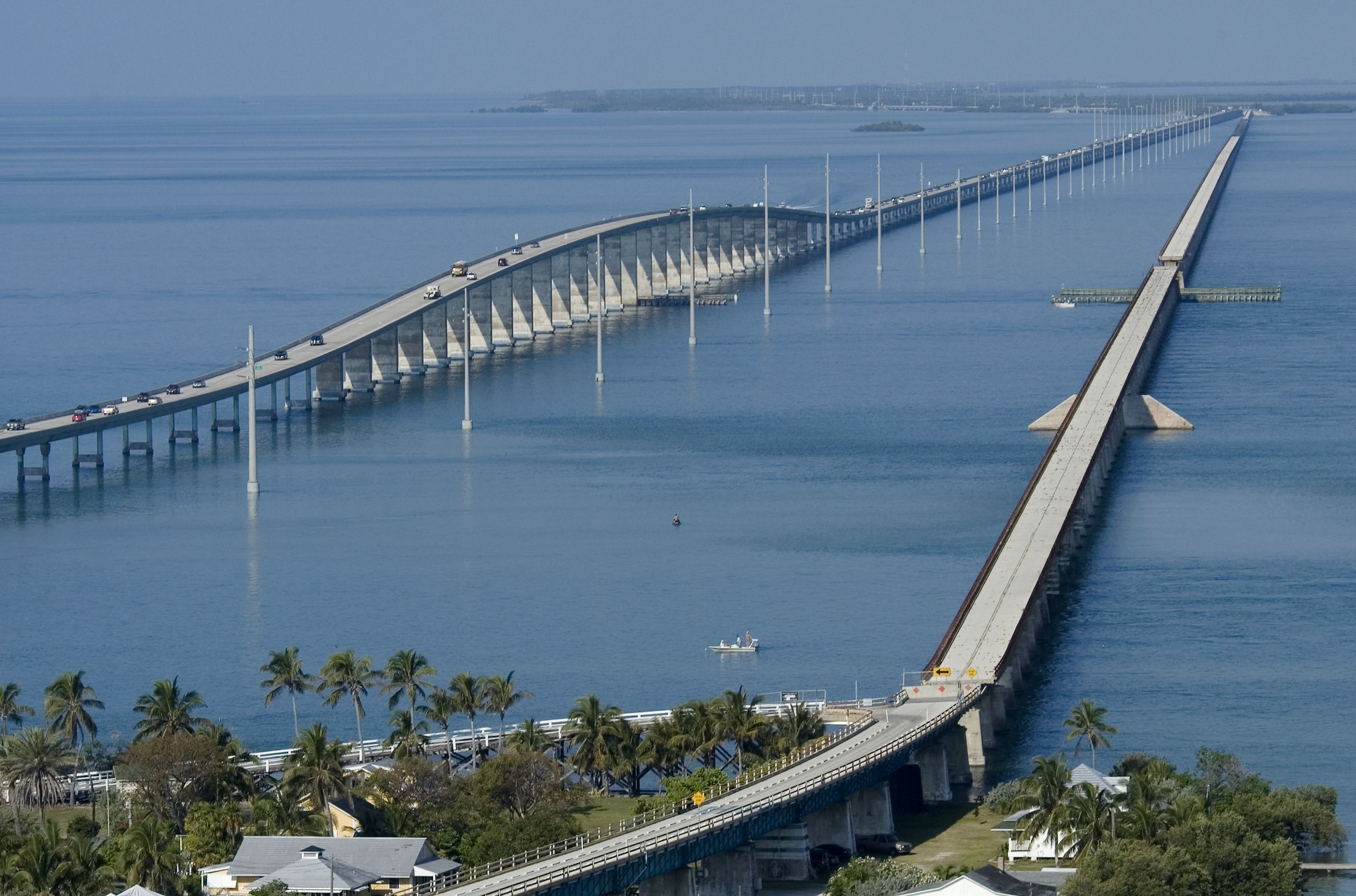 Aerial view of a long low bridge crossing calm blue ocean water, with palm trees and small buildings in foreground, clear sky